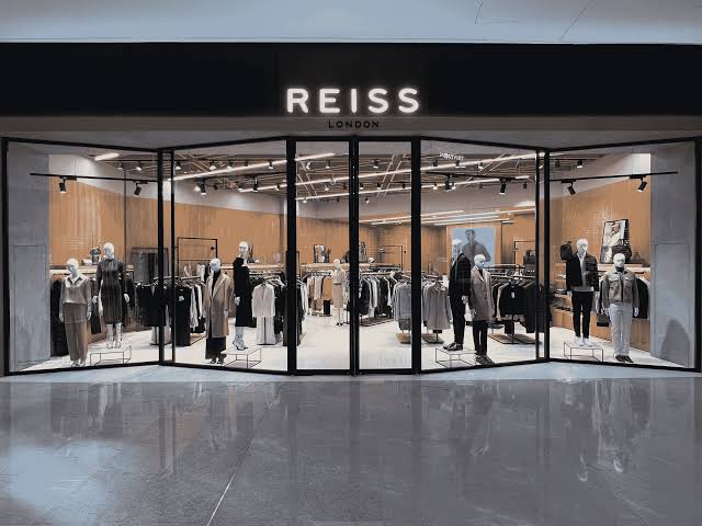 Reiss store frontage with slim black-framed glazing, timber-lined interior and track lighting